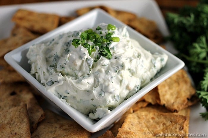 Garlic Herb Dip starts with a cream cheese base then is filled with fresh herbs. Pair this super easy dip recipe with crackers, chips, or veggies to make the perfect quick snack for your next party or game day. | www.persnicketyplates.com #dip #appetizer #easyrecipe #partyfood #garlic close up of garlic herb dip in square bowl