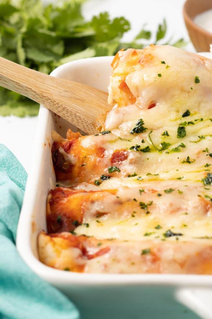 wooden spoon lifting an enchilada from a baking dish.