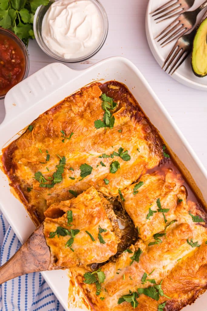 wooden spoon lifting an enchilada from a casserole dish.