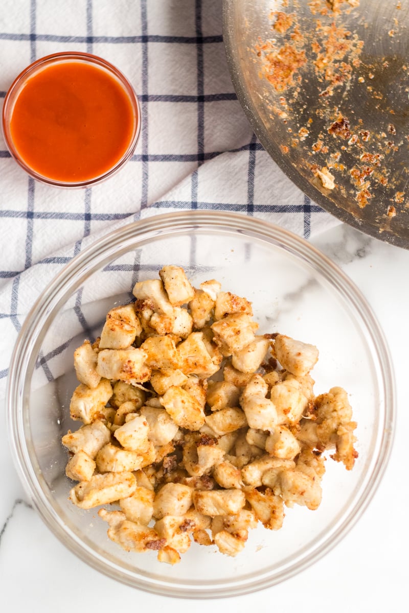 If you like the Buffalo Ranch Chicken Wrap from Buffalo Wild Wings, you'll love this lightened up homemade version! Lightly breaded buffalo chicken pieces wrapped up in a soft tortilla with ranch dressing, shredded lettuce, cheese, and tomatoes make a great dinner or the perfect game day meal! | www.persnicketyplates.com bowl of chicken with wing sauce for buffalo ranch chicken wraps