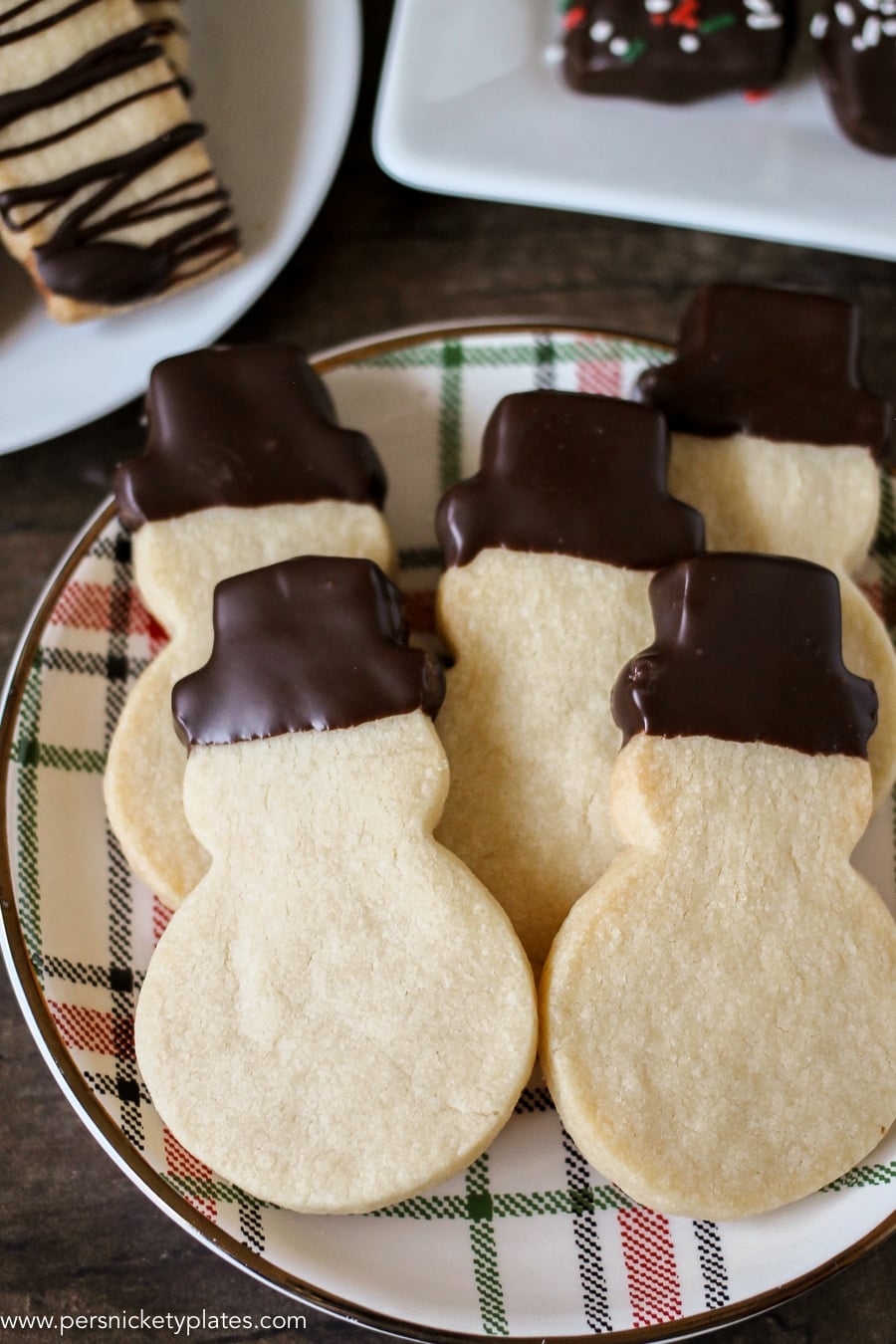 Chocolate Dipped Shortbread Cookies are a simple, from-scratch, shortbread cookie dipped in dark chocolate that's perfect for dunking in milk, coffee, hot chocolate...Santa won't be disappointed with these shortbread Christmas cookies! | www.persnicketyplates.com #christmascookies #shortbread #easyrecipe #baking #cookies #chocolate #sprinkles plate of snowman shaped shortbread cookies with chocolate dipped hats