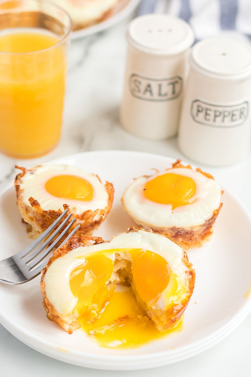 Bird's Nest Breakfast Cups are a fun way to eat eggs and hash browns for breakfast. These baked egg cups combine hash browns and cheese to form a "nest" and are topped with an egg. | www.persnicketyplates.com plate of egg in hash brown cups with cracked yolk spilling out