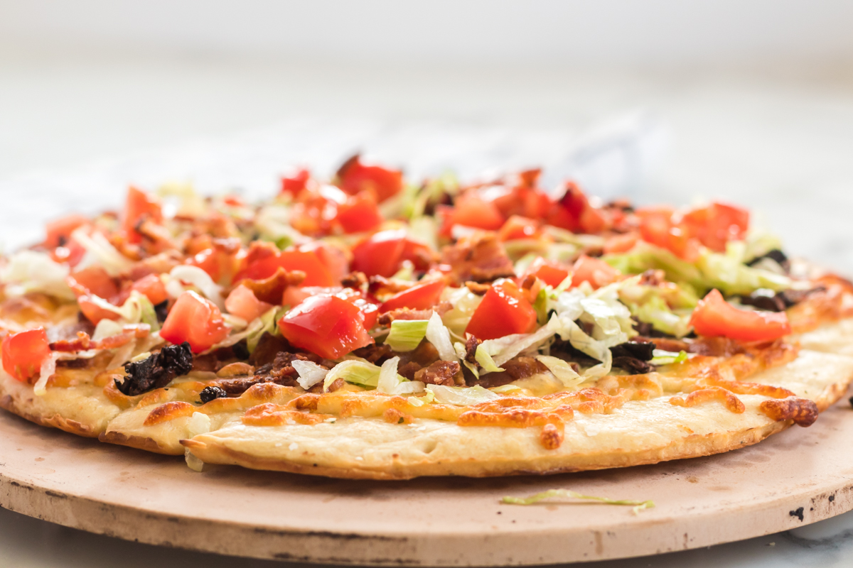 This BLT Pizza is semi-homemade and topped with cheese, bacon, lettuce, and tomatoes then drizzled with ranch dressing - a super easy meal for pizza night at home! | www.persnicketyplates.com side view of blt pizza on pizza stone