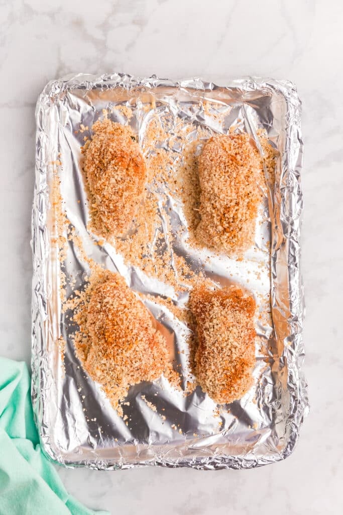 Four breaded stuffed chicken breasts on a foil-lined baking sheet.