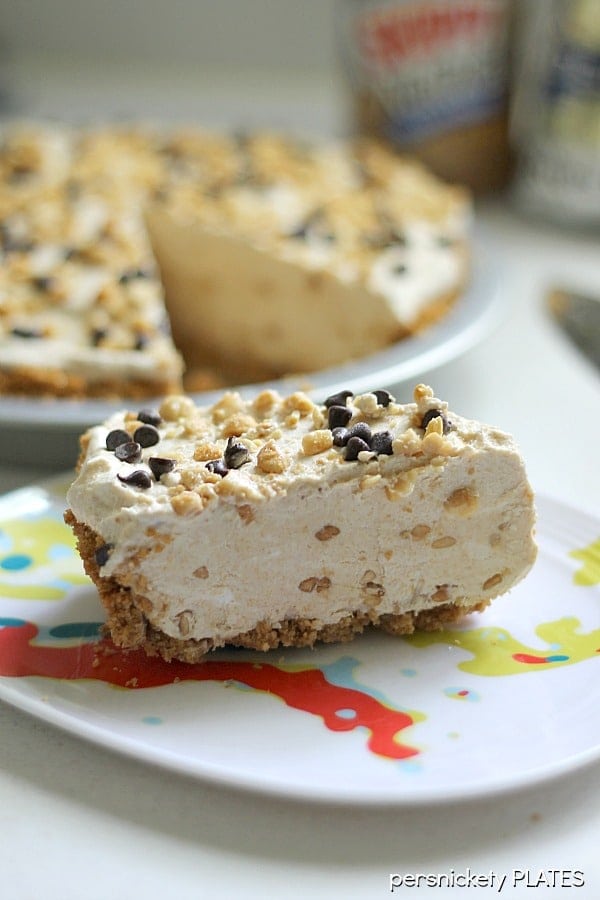 slice of frozen peanut butter pie on plate with rest of pie in background slice of peanut butter pie topped with peanuts and mini chocolate chips.