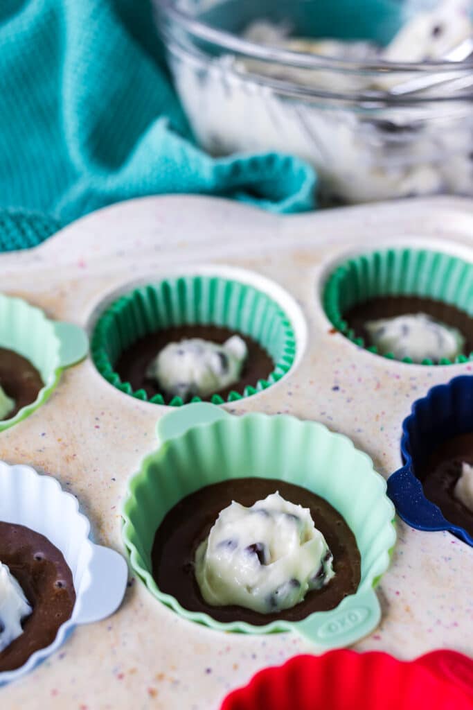 A muffin pan with chocolate batter and a dollop of cheesecake filling in each cup.