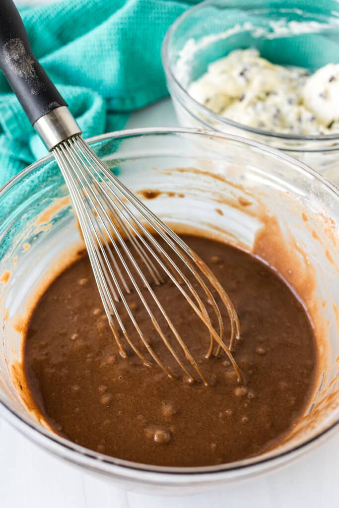 A glass bowl of chocolate cupcake batter with a metal whisk inside.