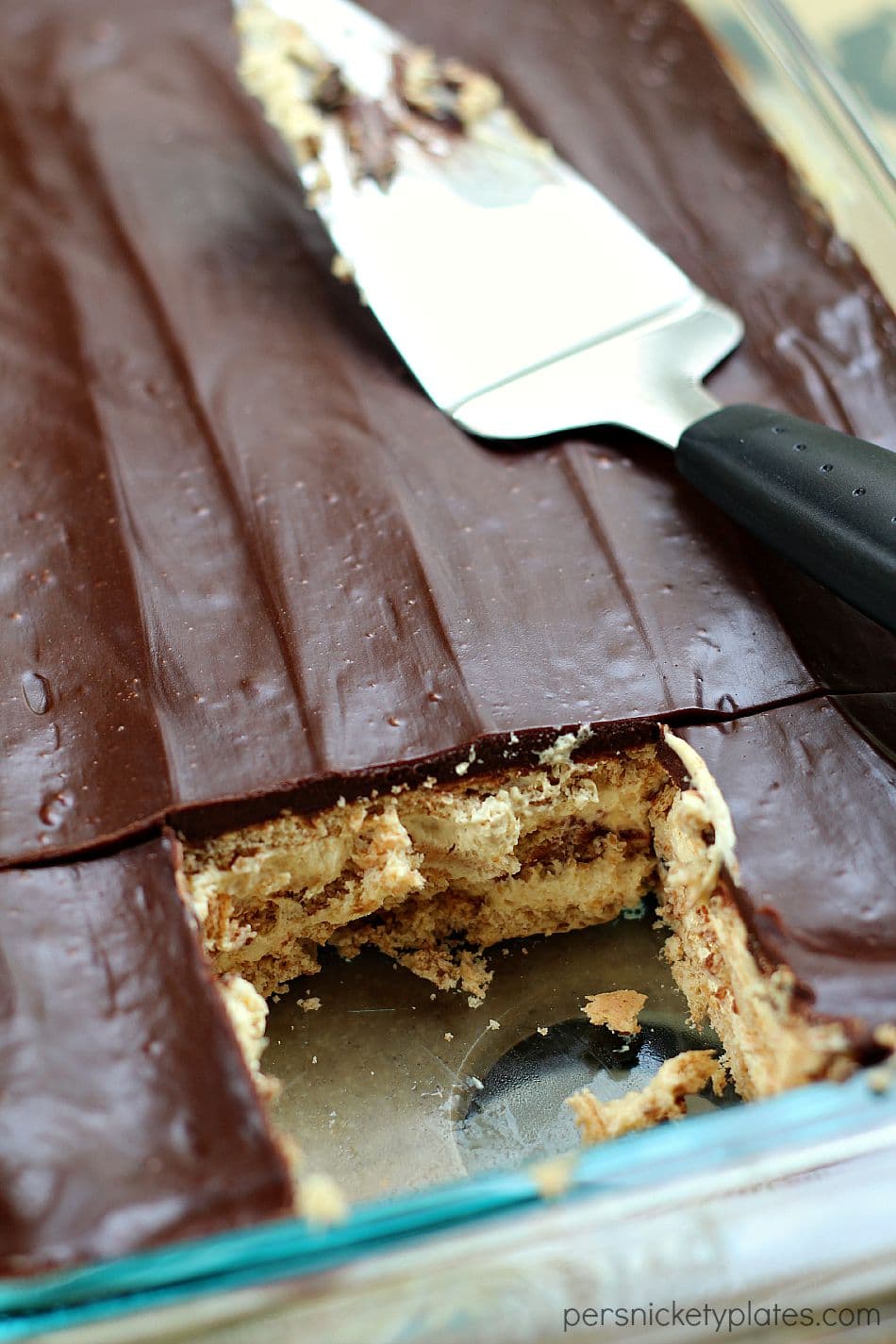 This no bake Chocolate and Peanut Butter Eclair Dessert has layers of graham crackers, layers of peanut butter pudding, a sprinkling of mini chocolate chips, and a chocolate peanut butter frosting. It's a chocolate + peanut butter lovers dream. | www.persnicketyplates.com pan of chocolate peanut butter eclair dessert in baking dish with serving spoon