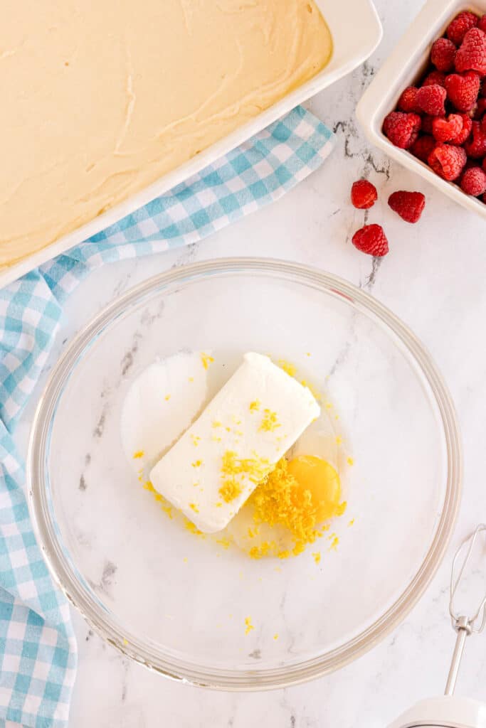 Glass bowl with cream cheese, sugar, egg, and lemon zest on a marble counter beside a white baking dish and a container of fresh raspberries.