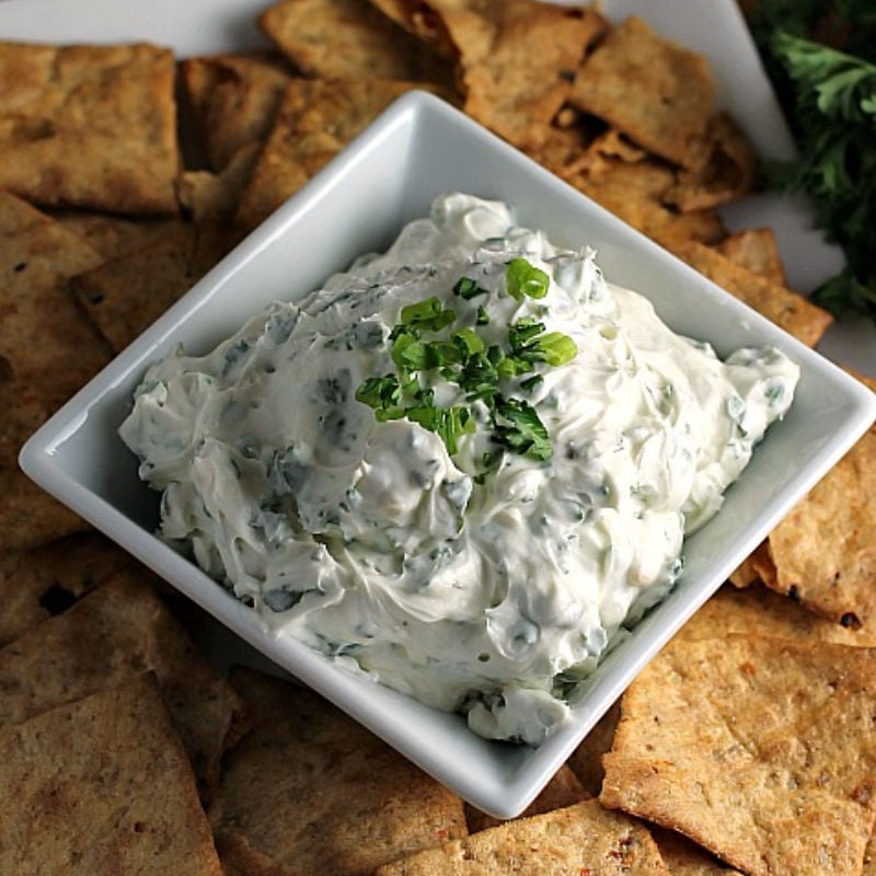 Garlic Herb Dip starts with a cream cheese base then is filled with fresh herbs. Pair this super easy dip recipe with crackers, chips, or veggies to make the perfect quick snack for your next party or game day. | www.persnicketyplates.com #dip #appetizer #easyrecipe #partyfood #garlic garlic herb dip in square bowl surrounded by chips