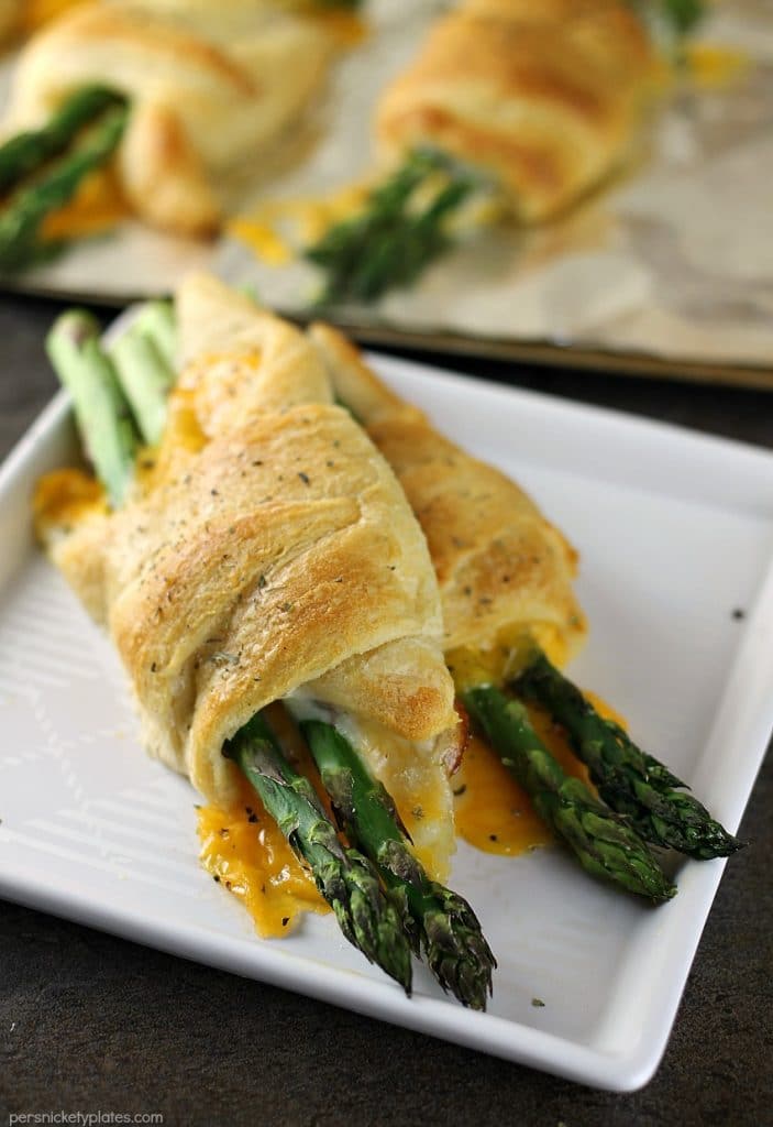 Cheesy Asparagus Puffs are a quick appetizer or side dish. Fresh asparagus wrapped up with provolone and cheddar cheeses and baked in a crescent roll - easy, cheesy, and delicious! | www.persnicketyplates.com #cheese #cheesy #appetizer #asparagus #vegetarian two cheesy asparagus puffs on a white plate