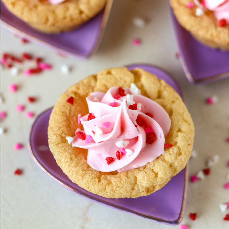 sugar cookie cup with pink frosting & heart sprinkles on purple plate Valentine Sugar Cookie Cups are a simple, semi-homemade treat that you can whip up in no time! A sugar cookie base filled with pink frosting and topped with Valentine sprinkles. | www.persnicketyplates.com