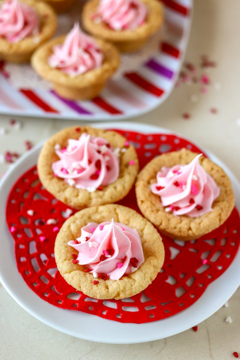 plate of valentine sugar cookie cups on red doily Valentine Sugar Cookie Cups are a simple, semi-homemade treat that you can whip up in no time! A sugar cookie base filled with pink frosting and topped with Valentine sprinkles. | www.persnicketyplates.com