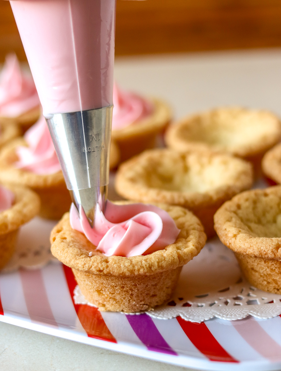pink frosting being piped into valentine sugar cookie cups Valentine Sugar Cookie Cups are a simple, semi-homemade treat that you can whip up in no time! A sugar cookie base filled with pink frosting and topped with Valentine sprinkles. | www.persnicketyplates.com