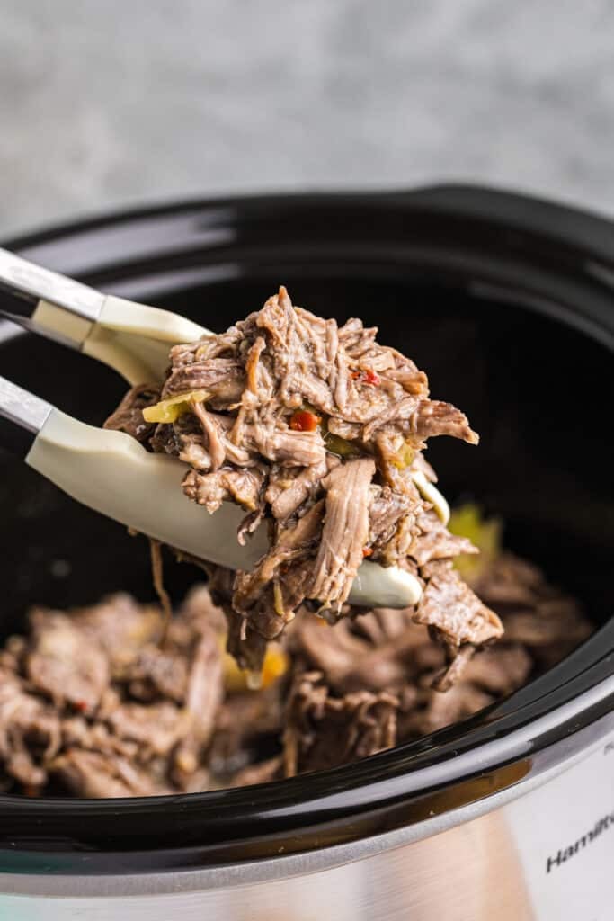 tongs lifting shredded beef from a crockpot.