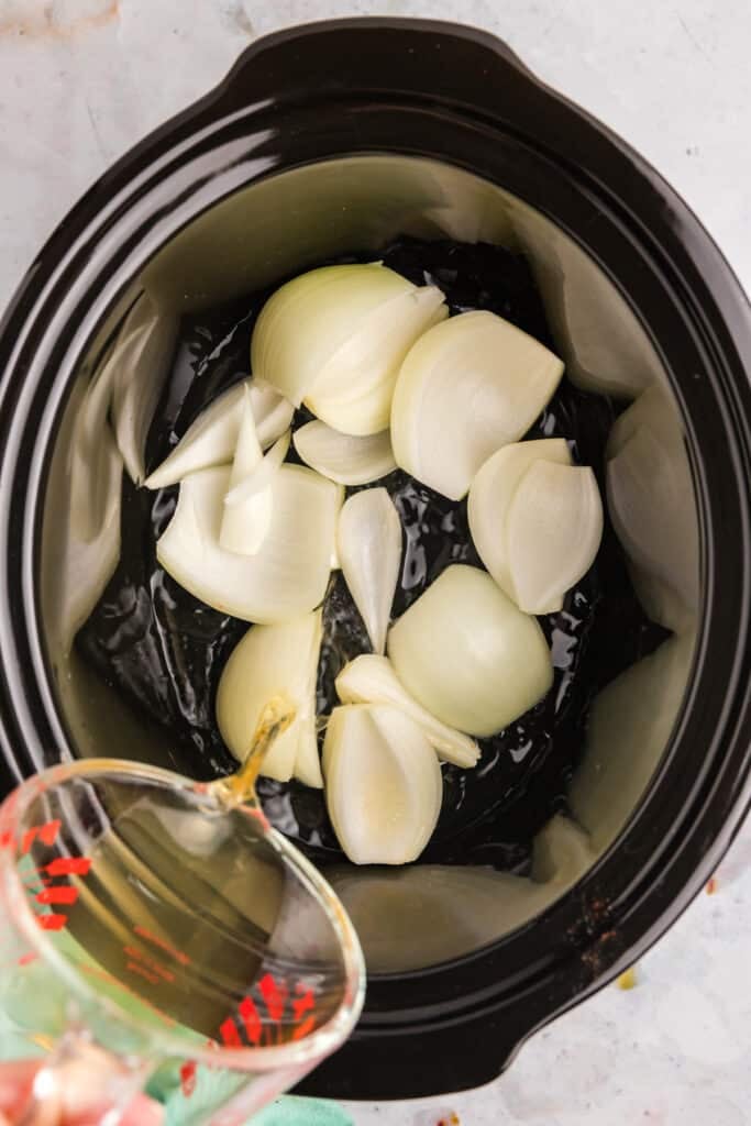 Quartered onions in a crockpot with broth being poured.