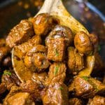 wooden spoon lifting a scoop of mushroom steak bites from a slow cooker.