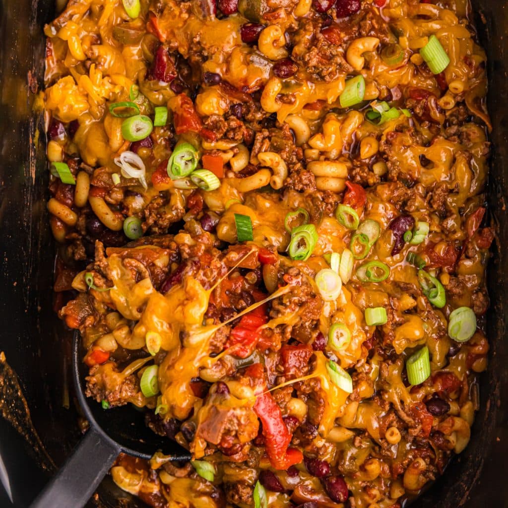 overhead shot of a spoon scooping chili mac from a slow cooker.
