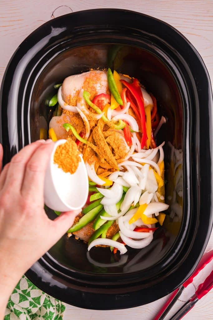 hand sprinkling taco seasoning onto peppers, onions, and chicken in a slow cooker.