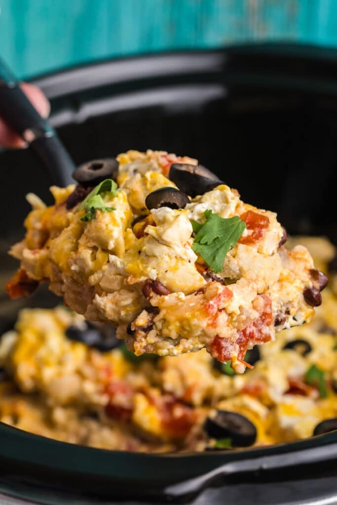 spoon lifting a portion of enchilada casserole from a slow cooker.