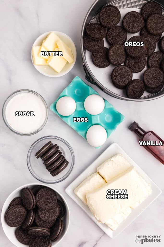 An overhead view of labeled ingredients for cookies and cream cheesecake, including butter, sugar, eggs, vanilla, cream cheese, and Oreos, arranged on a marble countertop.