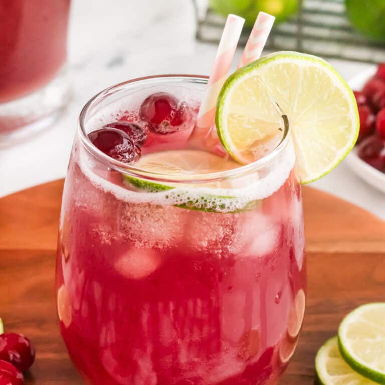 A glass of cranberry punch with lime slice, cranberries, and pink-striped straws on a wooden tray.