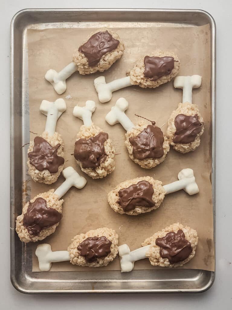 A tray of Rice Krispies treats shaped like turkey legs with white candy bones drizzled with chocolate on parchment paper.