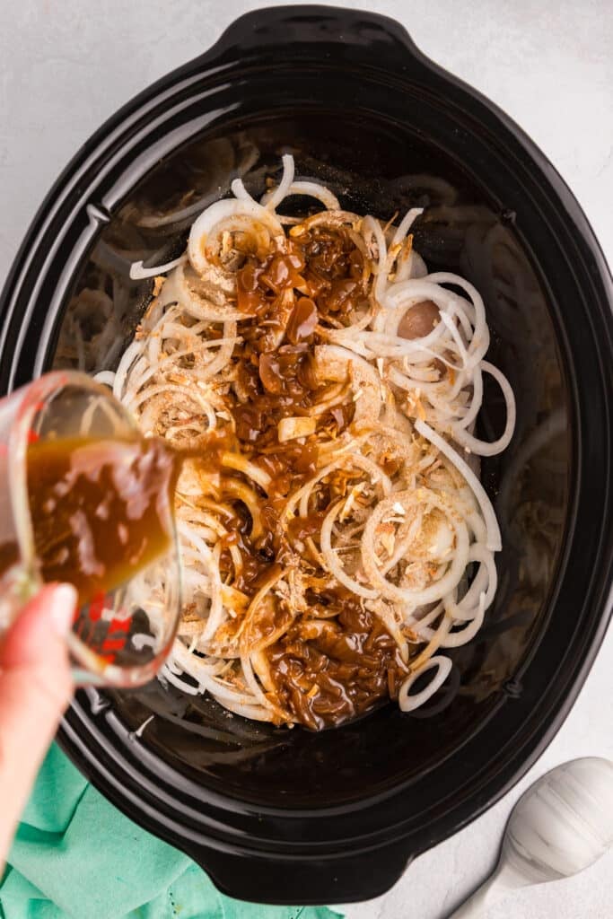 Pouring brown sauce over sliced onions and chicken in a slow cooker