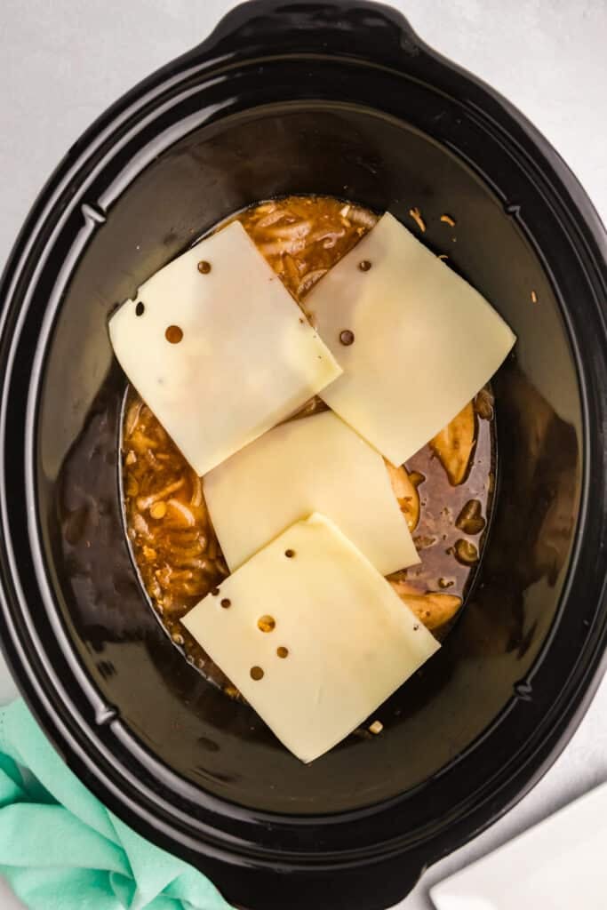 Swiss cheese slices layered on top of cooked chicken in a slow cooker.