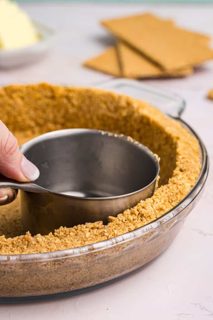 A hand pressing graham cracker crumbs into a glass pie dish with a measuring cup, forming a uniform crust along the sides and bottom.