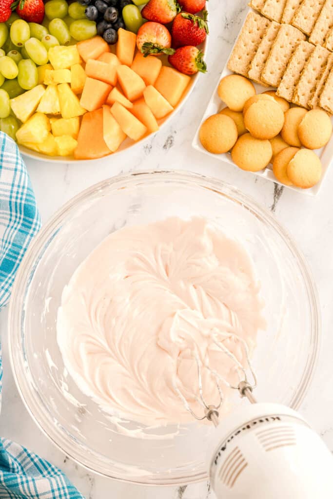 A glass bowl of creamy pink fruit dip being mixed with a hand mixer.
