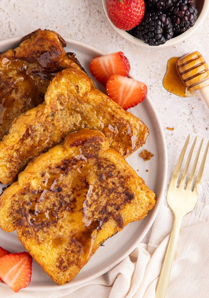A plate of golden French toast drizzled with syrup, served with sliced strawberries and a gold fork.