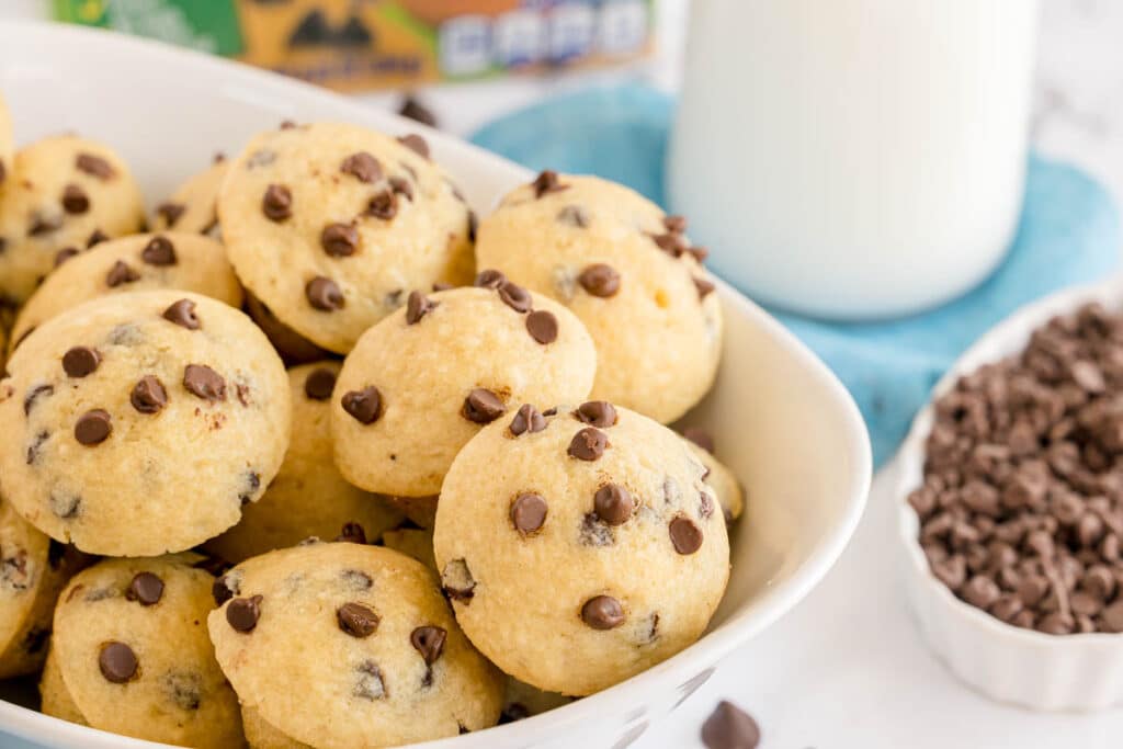 Bowl full of golden mini chocolate chip muffins with a glass of milk and chocolate chips in the background.