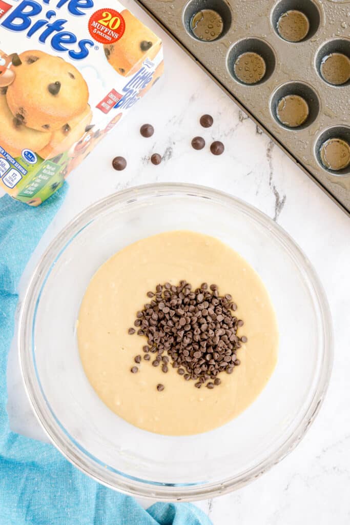 Mini chocolate chips piled on top of muffin batter in a glass bowl, with a muffin tin and box of Little Bites nearby.