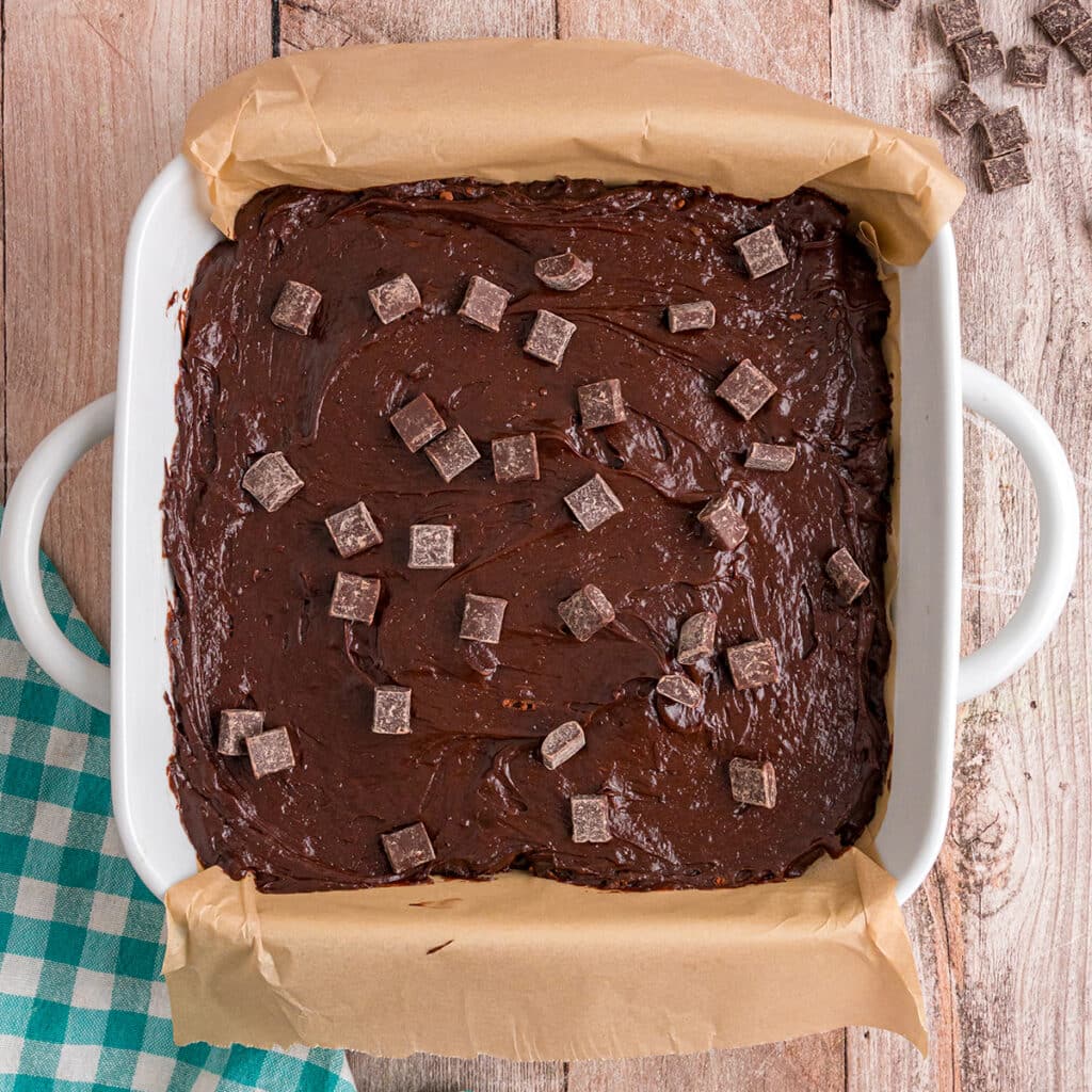Chocolate brownie batter spread in a parchment-lined white baking dish, topped with chocolate chunks.