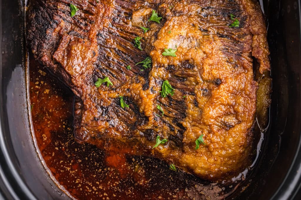 Cooked brisket in a slow cooker with visible juices and parsley.