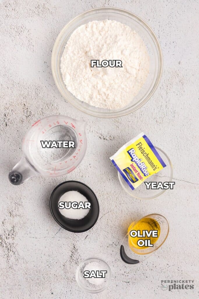 A top-down view of baking ingredients on a white surface, including flour, yeast, oil, and measuring tools.