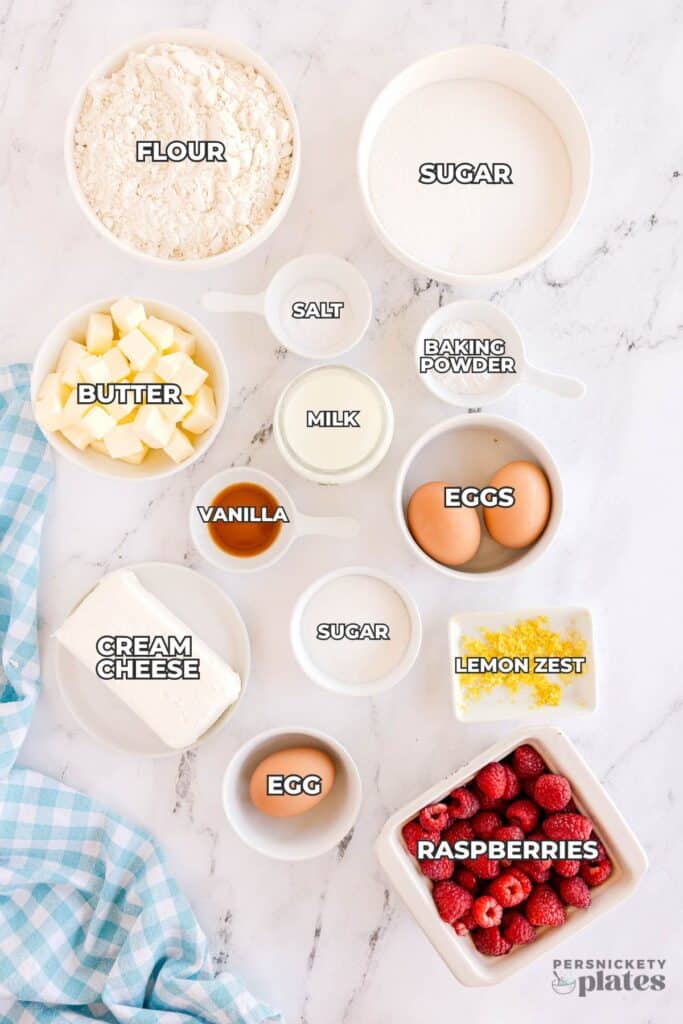 Overhead shot of labeled baking ingredients including flour, sugar, butter, cream cheese, eggs, raspberries, and lemon zest on a white countertop.