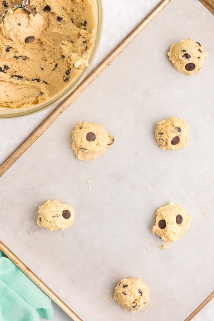 Scoops of cookie dough with chocolate chips arranged on a parchment-lined baking sheet next to a mixing bowl.