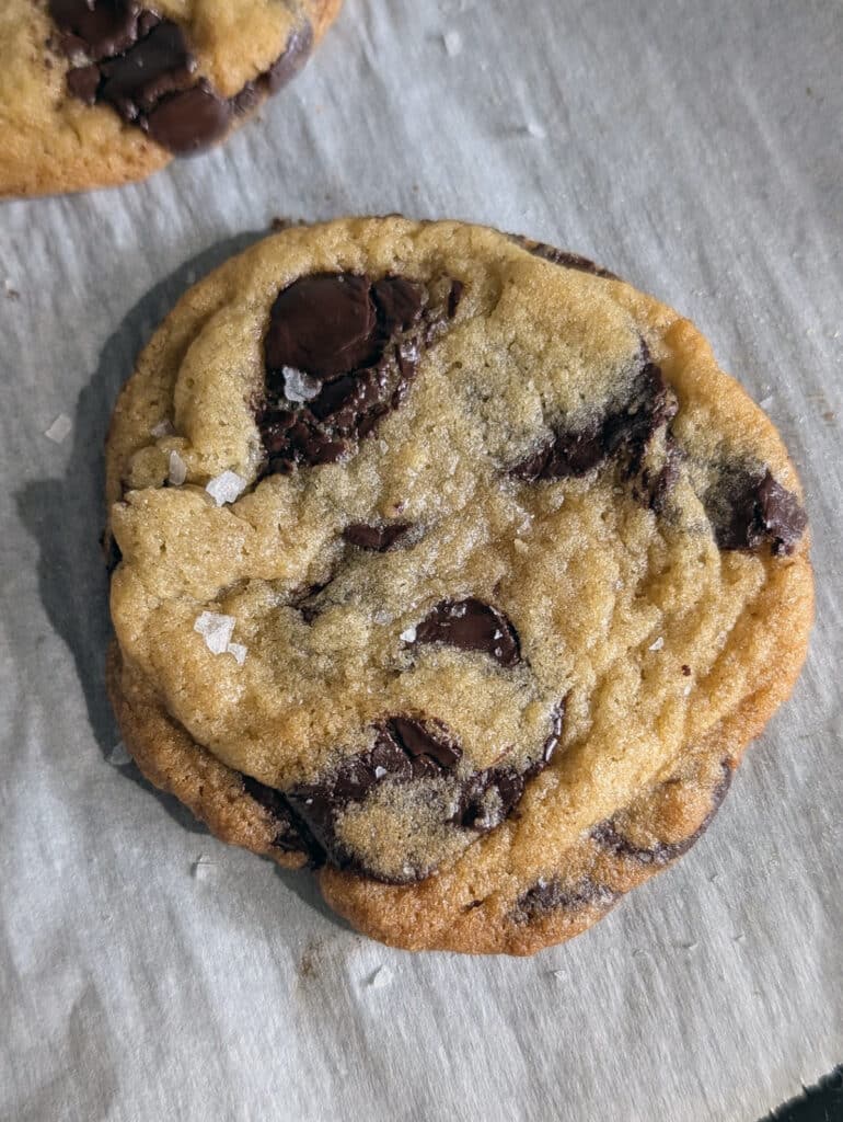 A single chocolate chip cookie on parchment paper with visible flaky sea salt and melted chocolate chunks.