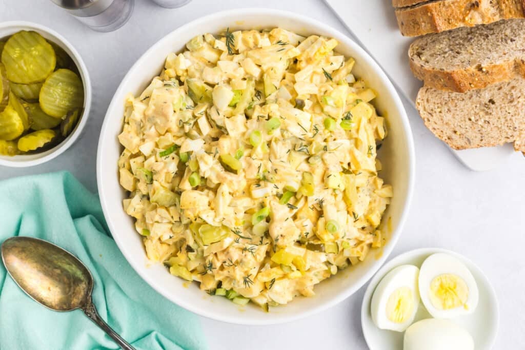 Overhead view of dill pickle egg salad in a white bowl surrounded by ingredients.