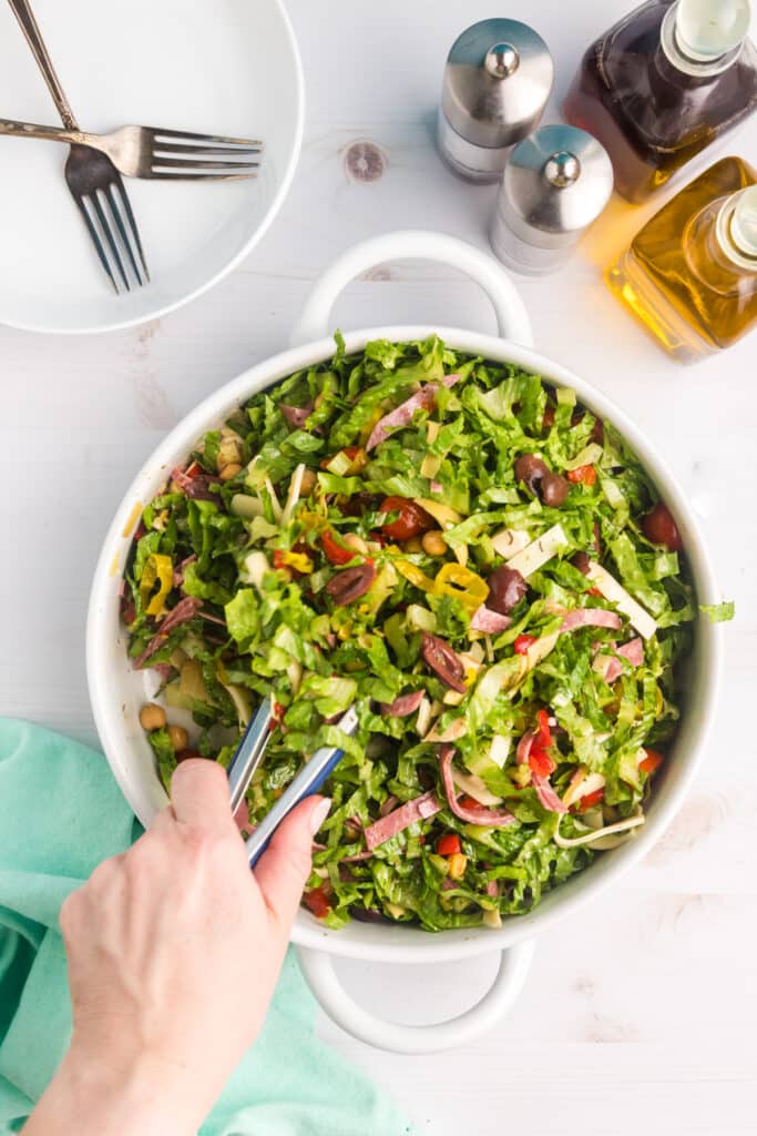A hand with metal tongs is mixing a large bowl of antipasto salad in a white bowl.