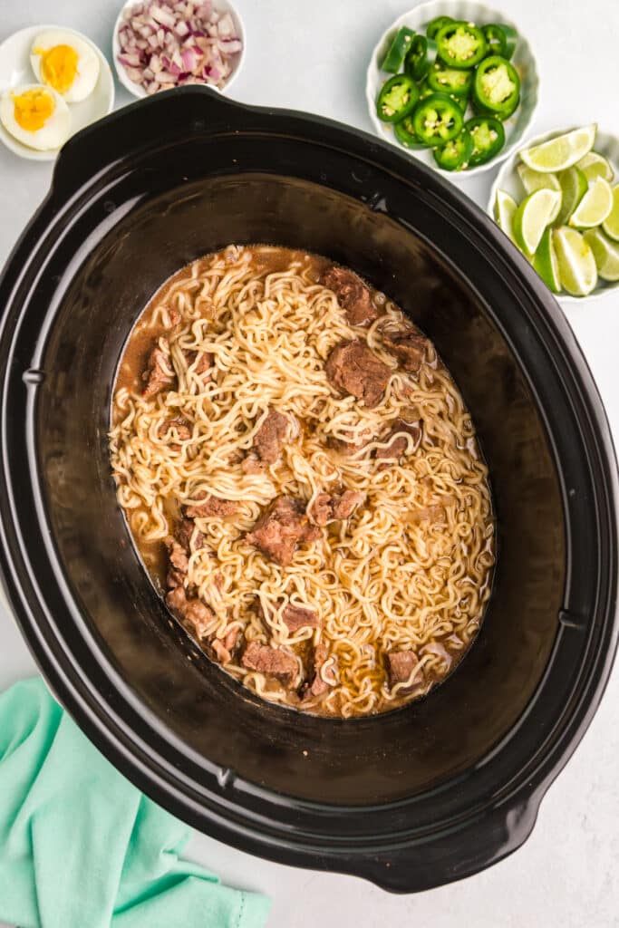 Slow cooker filled with cooked birria ramen next to bowls of toppings.