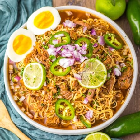 Close-up of birria ramen in a bowl with lime slices, soft eggs, and jalapeƱos.