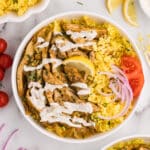 Bowl of yellow rice topped with sliced chicken shawarma, white sauce, and vegetables.