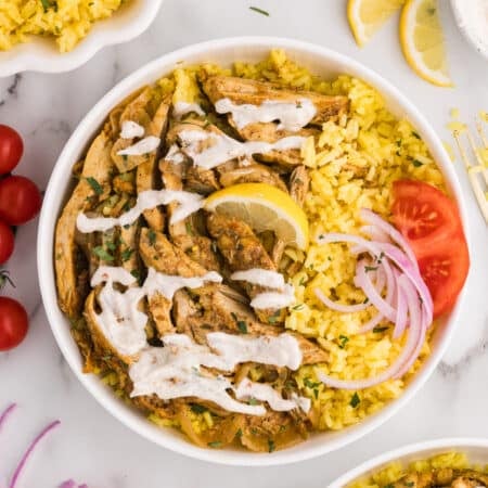 Bowl of yellow rice topped with sliced chicken shawarma, white sauce, and vegetables.