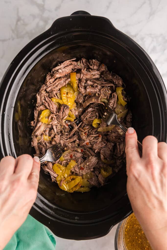 Hands using forks to shred beef and peppers inside a slow cooker.