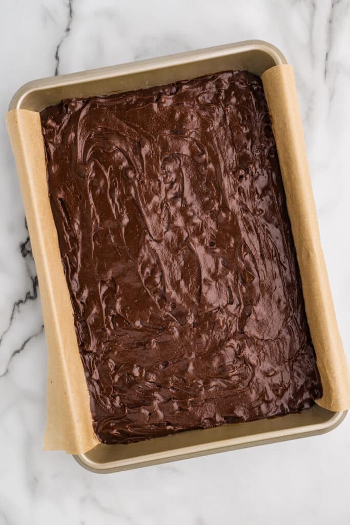 Brownie batter spread in a parchment-lined metal baking pan.