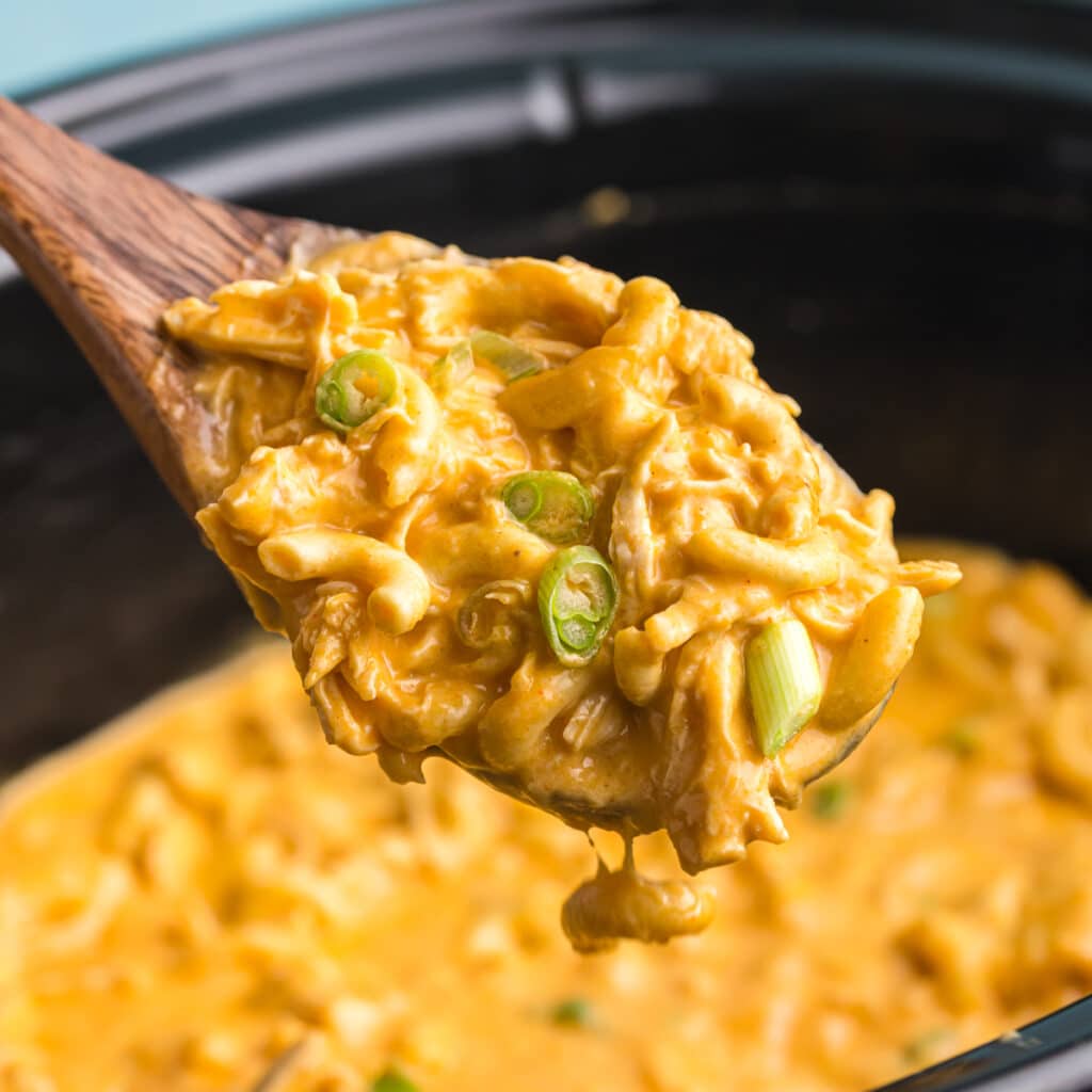 Wooden spoon lifting creamy buffalo chicken mac and cheese from slow cooker.