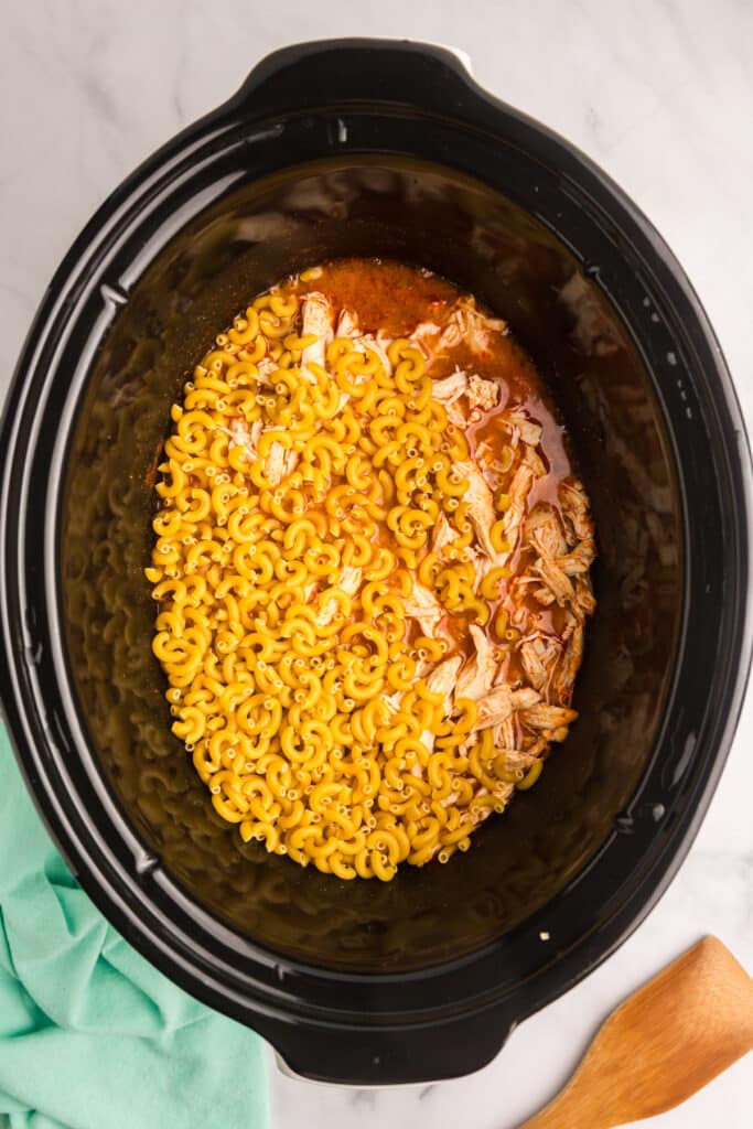 Uncooked elbow macaroni and shredded chicken in broth inside a slow cooker.